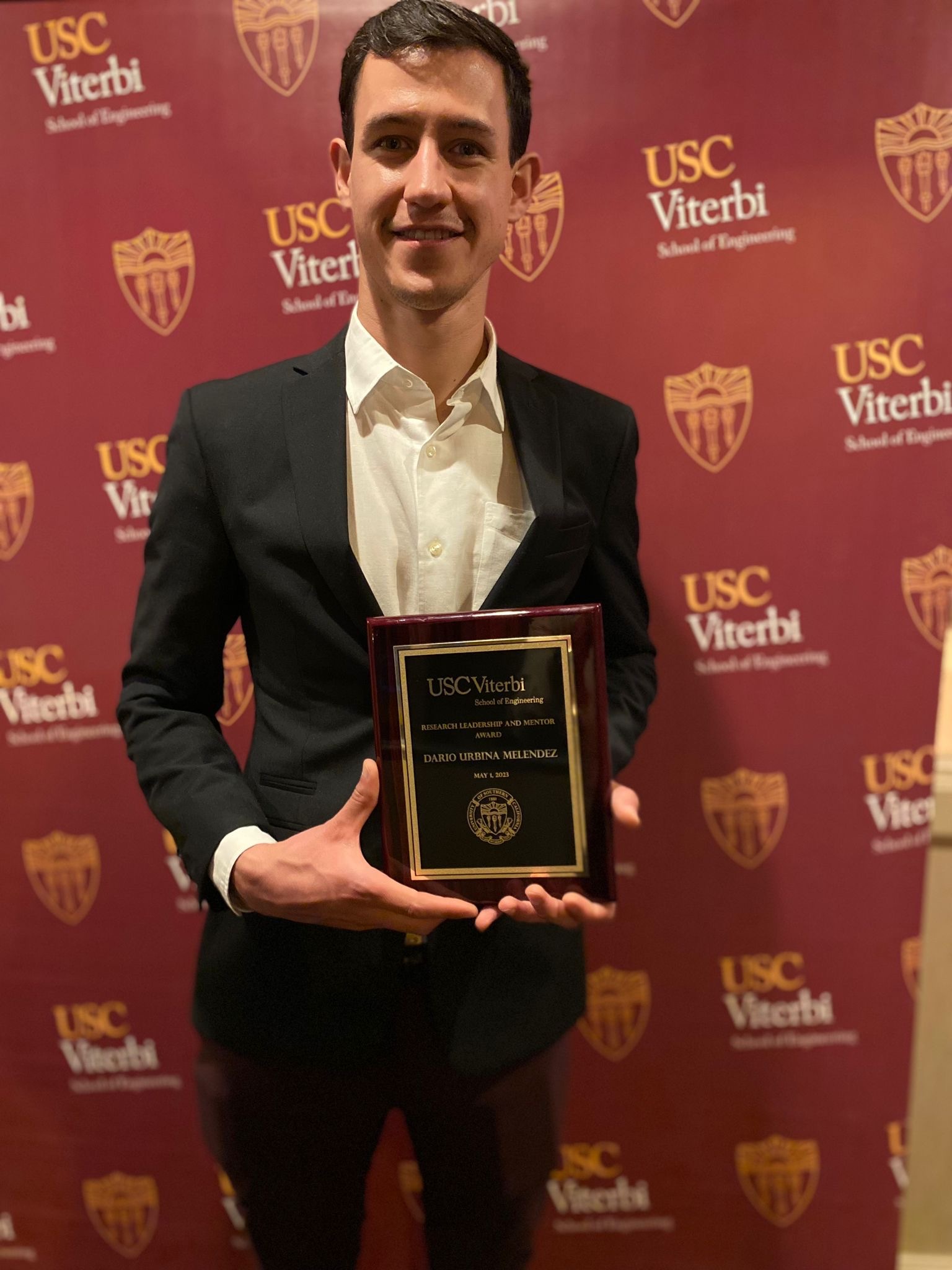 Dario smiles for the camera while holding the 2023 Viterbi Research Leadership and Mentor Award plaque. He is wearing a black suit with a white dress shirt and is standing in front of a red step-and-repeat with the USC Viterbi logo.
